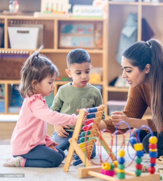 A female Kindergarten teacher of Middle Eastern decent, sits on the floor with students as they play with various toys and engage in different activities.  They are each dressed casually as they learn through their play.
